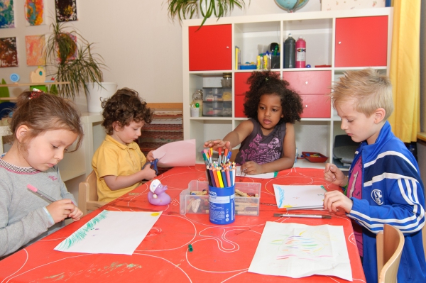 Vier Kinder sitzen am Tisch im Kreativbereich und malen mit Buntstiften und Filzstiften eigene kleine Kunstwerke. Ein Kind schneidet sein Papier.
