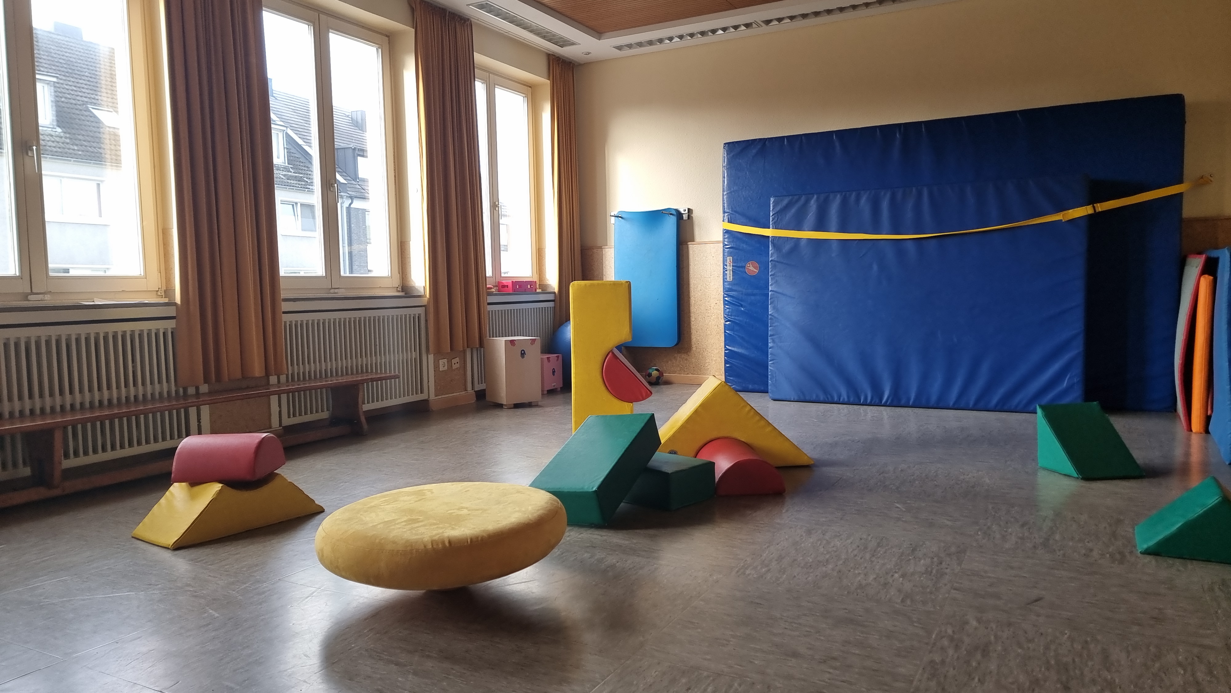 Hier ist den  Bewegungsraum mit den großen Bausteinelementen, einen gelben großen Balancierkreisel, verschiedenen Matten, eine Sportbank und im Hintergrund zwei Baff-Trommeln zu sehen.