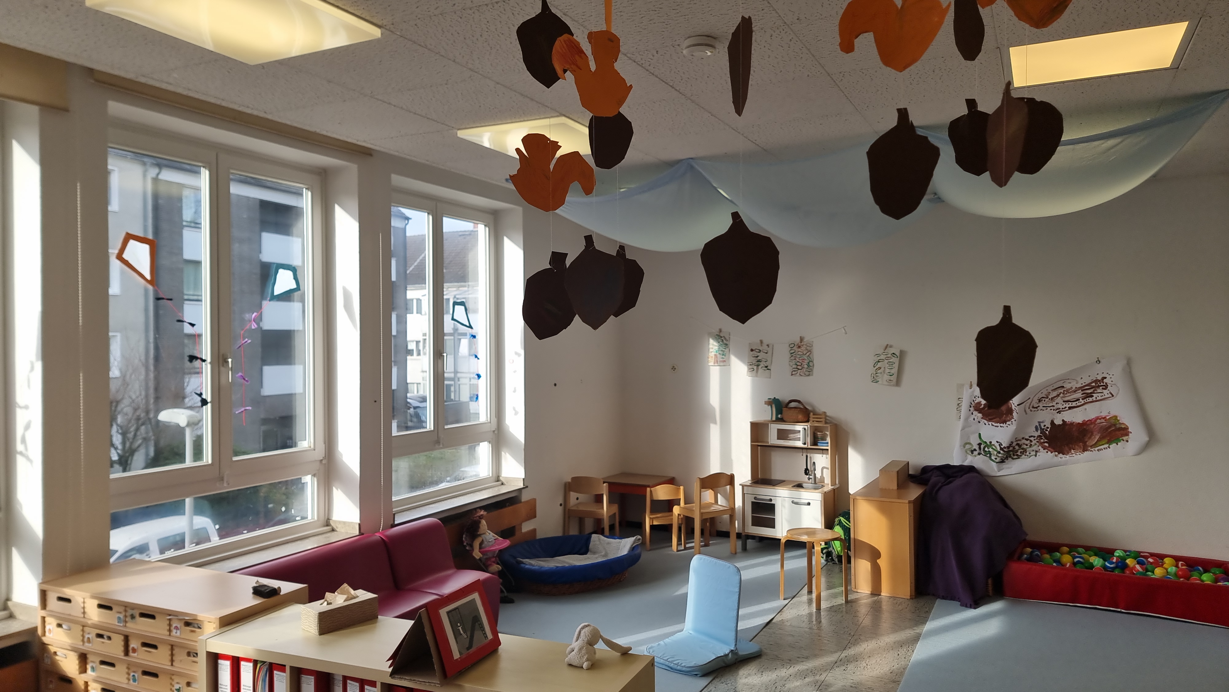 Auf diesem Bild ist der Rollenspielbereich mit einer Küche, einem Tisch und Stühlen, einem Sofa zu sehen. Daneben befindet sich ein Teppichbereich mit einem Becken mit Bällen. Im vorderen Ber