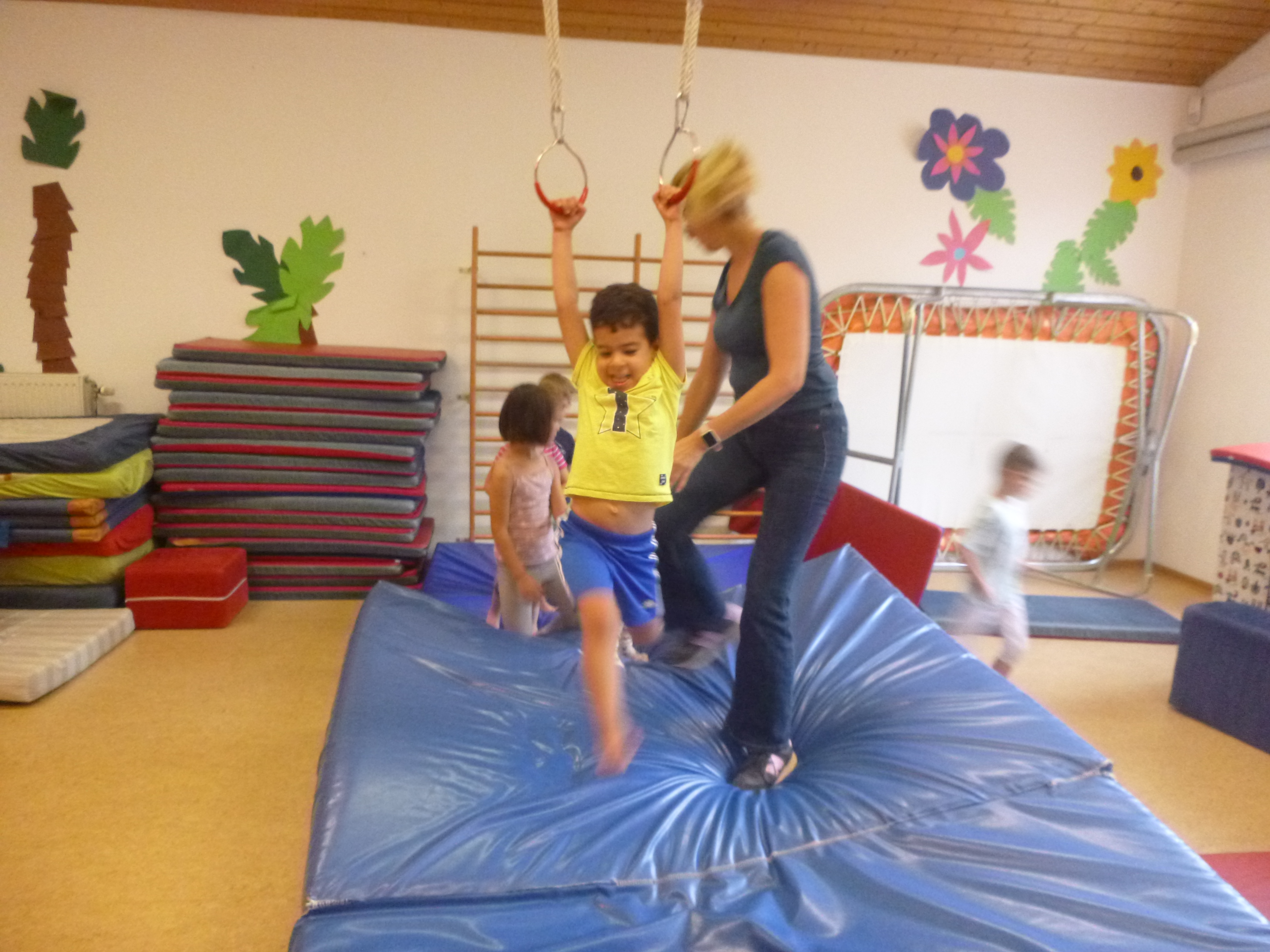 Es ist ein Bild aus der Turnhalle. Es ist eine große blaue Mitte mit Ringen zum Schwingen zu erkennen. Ein Kind hängt an den Ringen. Im Hintergrund sind noch weitere Kinder zu erkennen