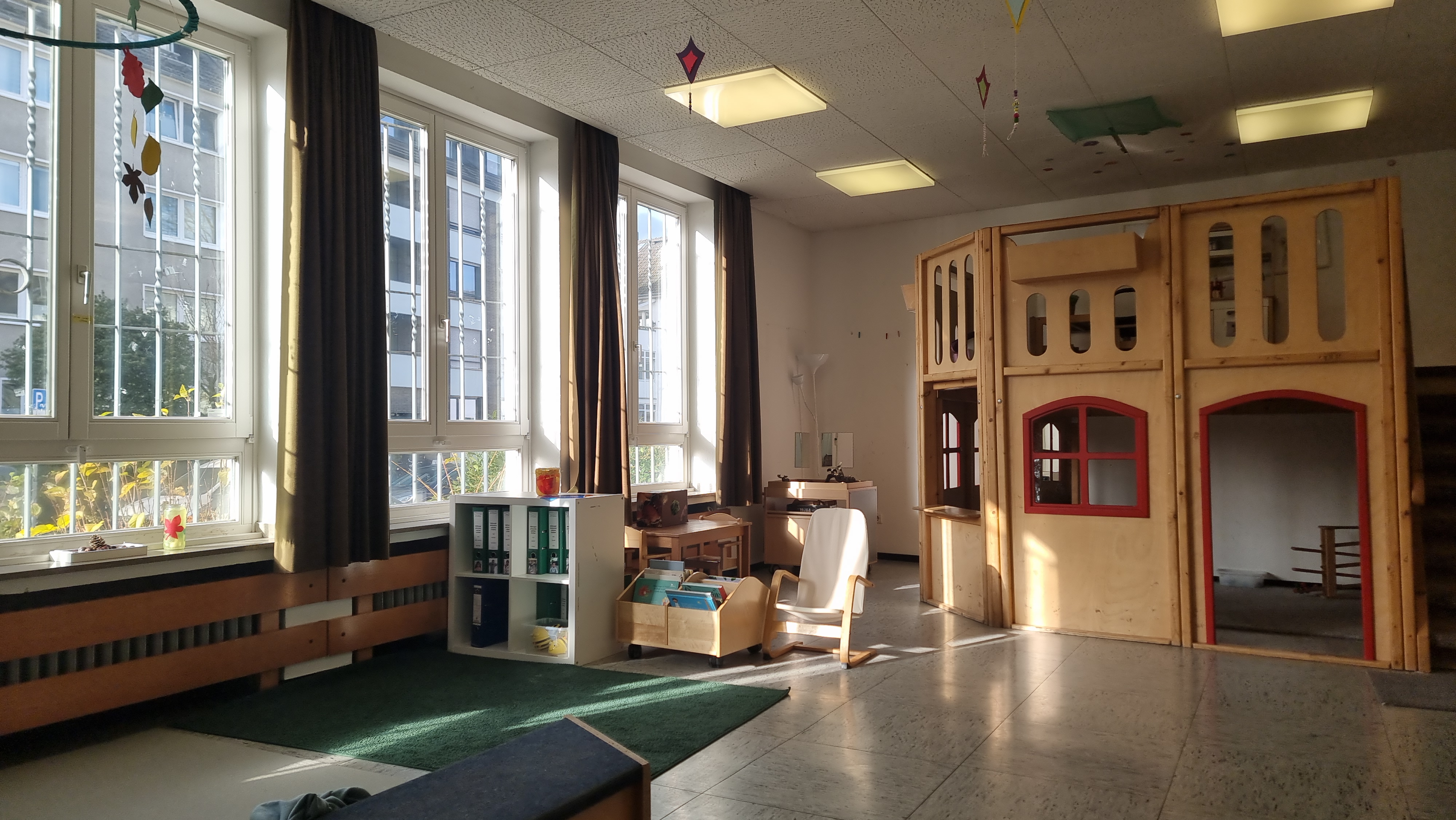 In diesem Gruppenraum ist ein Bereich mit Teppich , eine Bücherkiste, ein Forscherwagen und ein Spielhaus mit zwei Ebenen zu sehen. Der Teppichbereich ist ein Bereich, wo die Kinder sich ausr