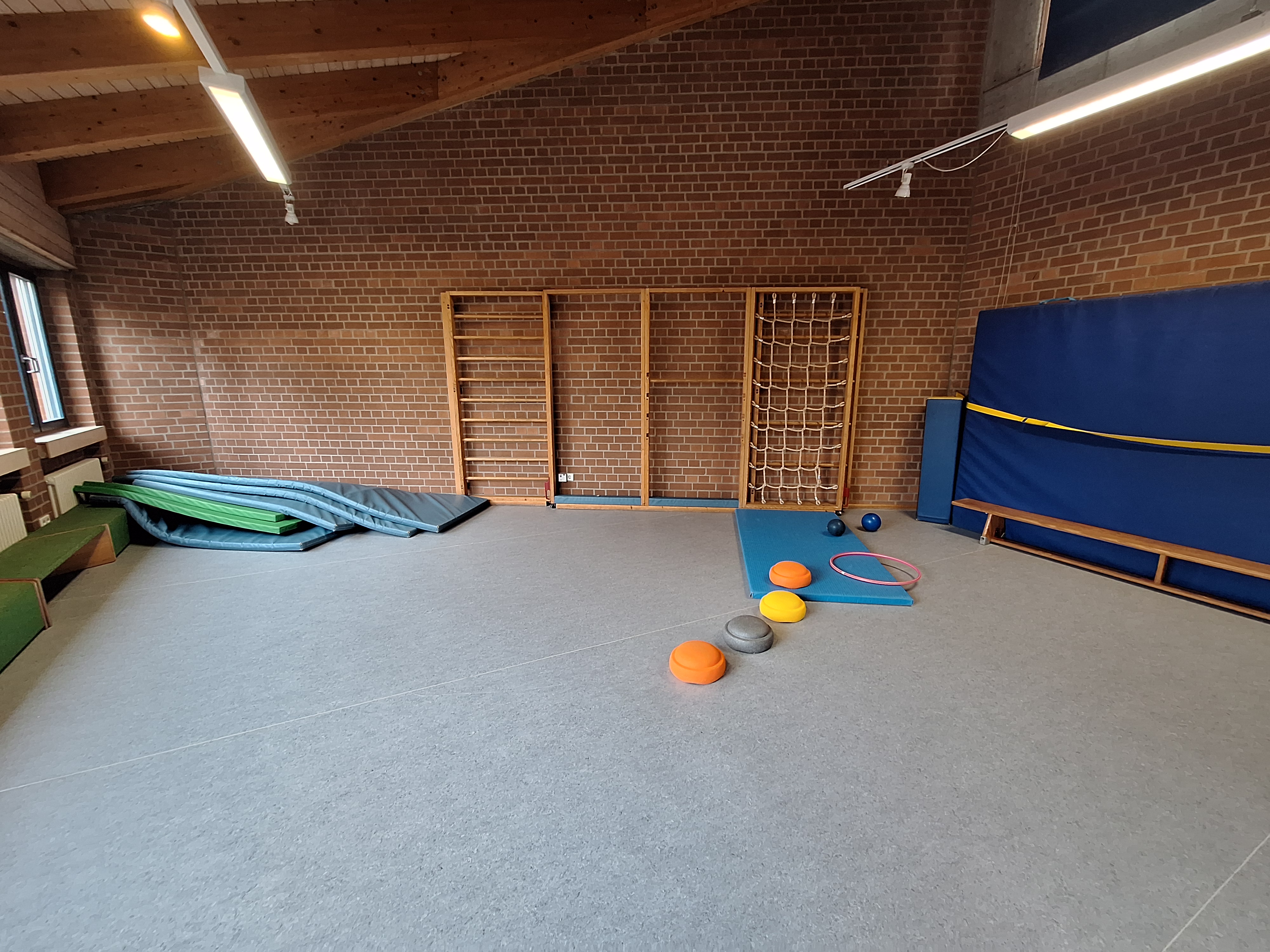 Mehrzweckraum der KiTa mit großer freier Fläche. An der Wand befindet sich ein Turnelement wie Sprossenwand und Kletternetz. Der Raum bietet viel Platz für Spielen, Bewegung und motorische Üb