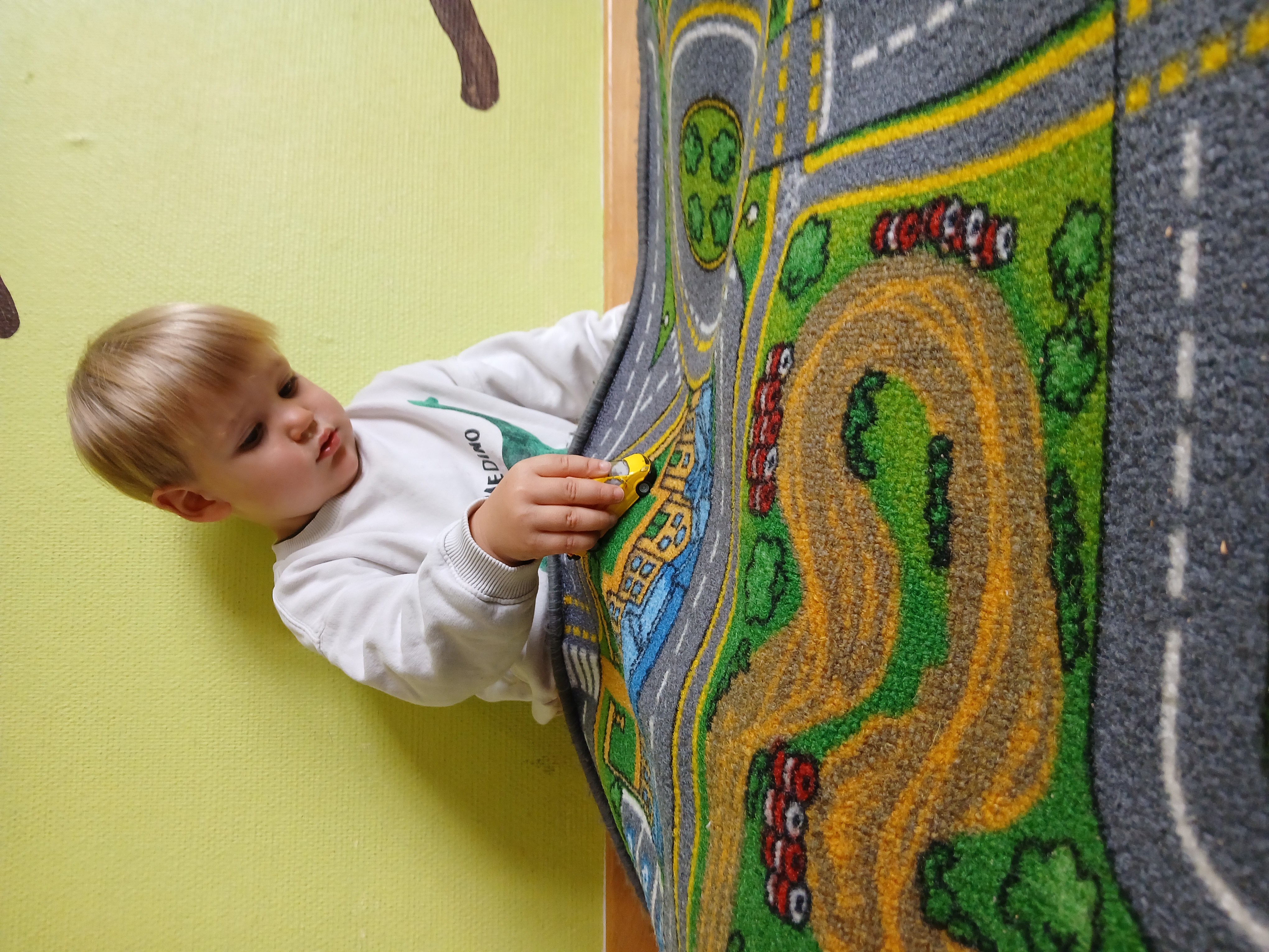 Ein Kind sitzt auf dem Boden und hat einen Straßenteppich auf den Knien. Darauf fährt er mit einem Spielzeugauto.