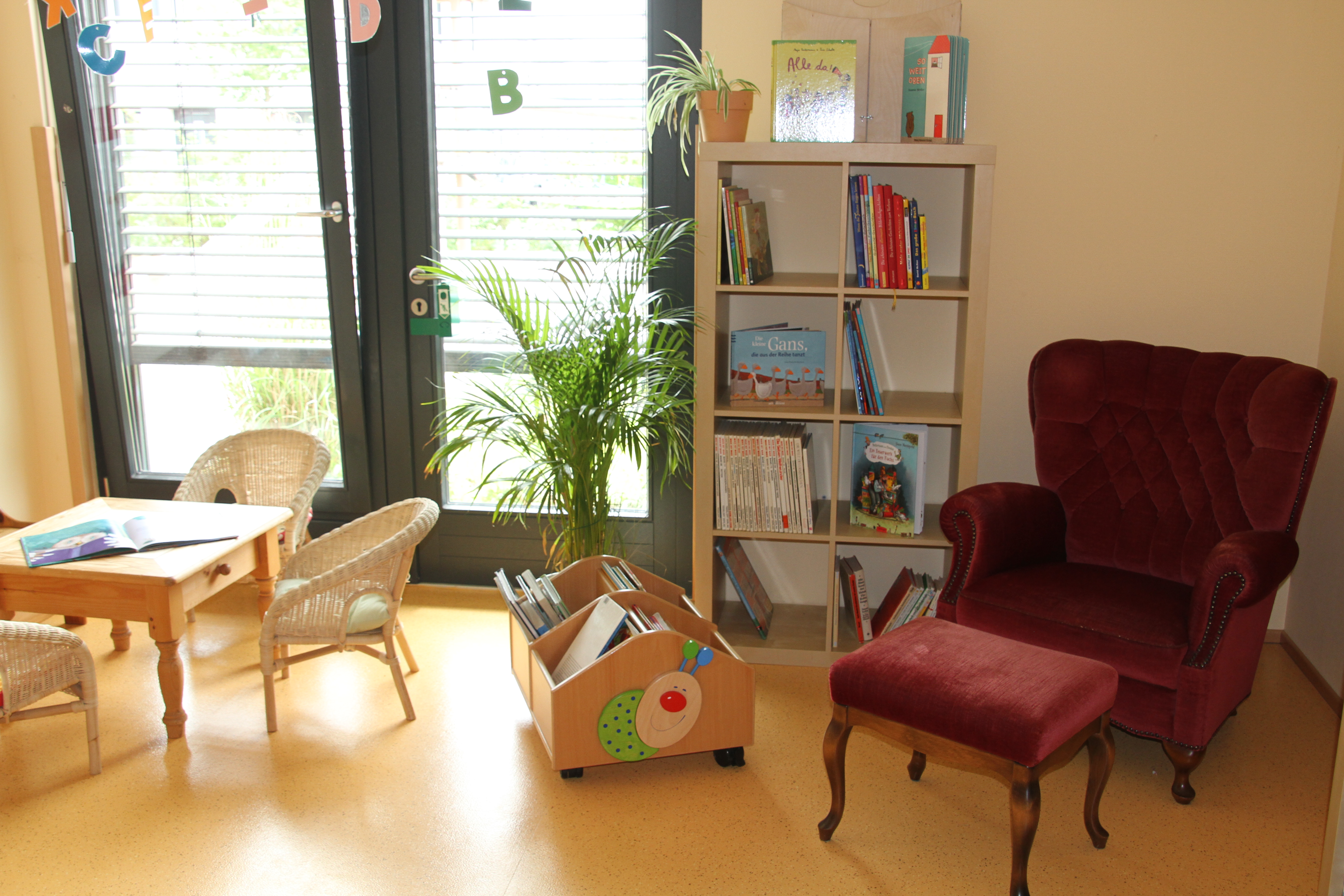 Das Bild zeigt einen hellen Raum mit tiefgezogenen Fensterelementen, in dem ein mit dunkelrotem Samt bezogener Ohrensessel steht, daneben ein Regal mit vielen Bilderbüchern. Vor dem Fenster s