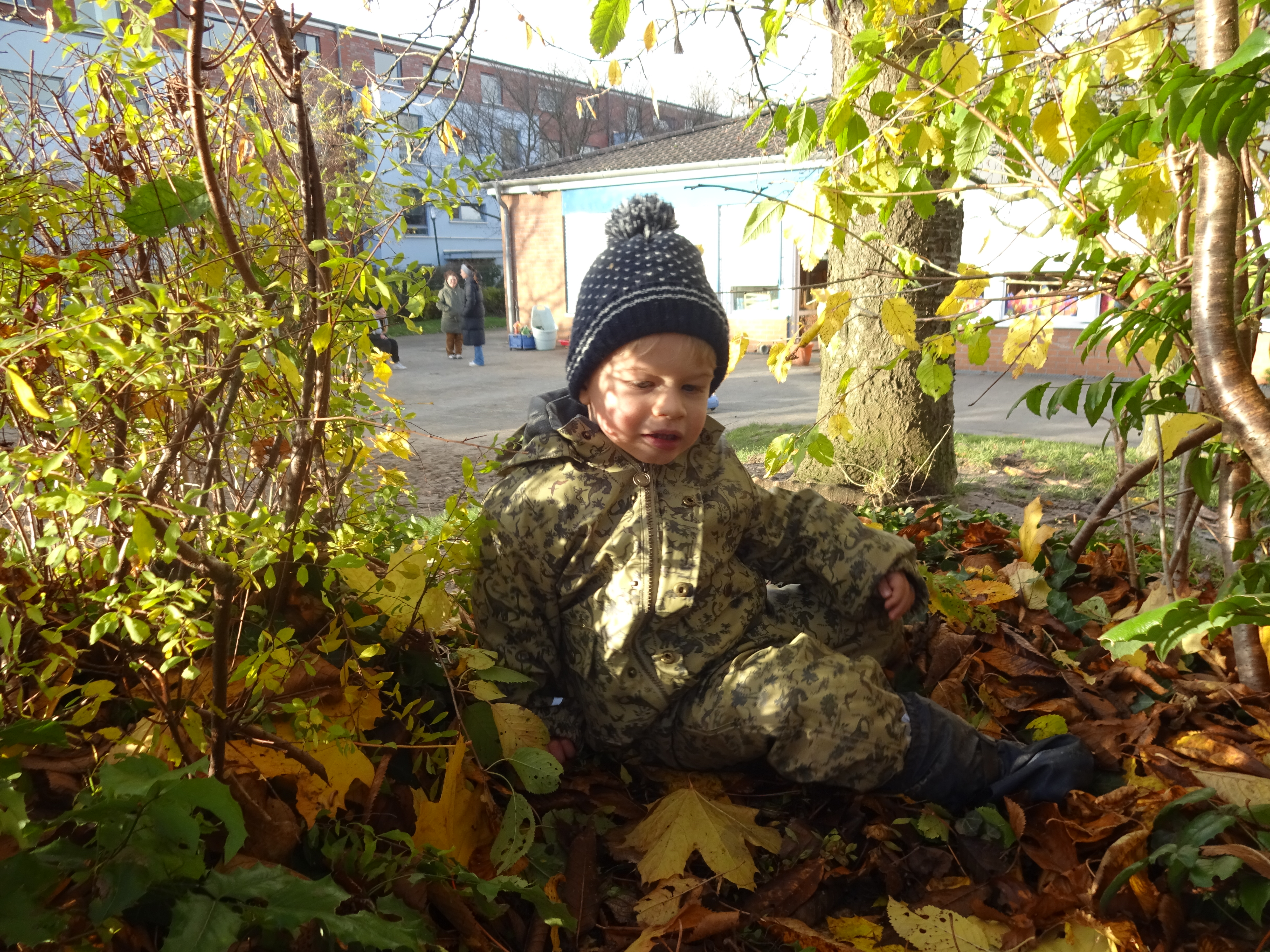 Ein Junge sitzt mit Mütze und im Matschanzug zwischen den Bäumen im Herbstlaub. Die Sonne scheint ihm ins Gesicht