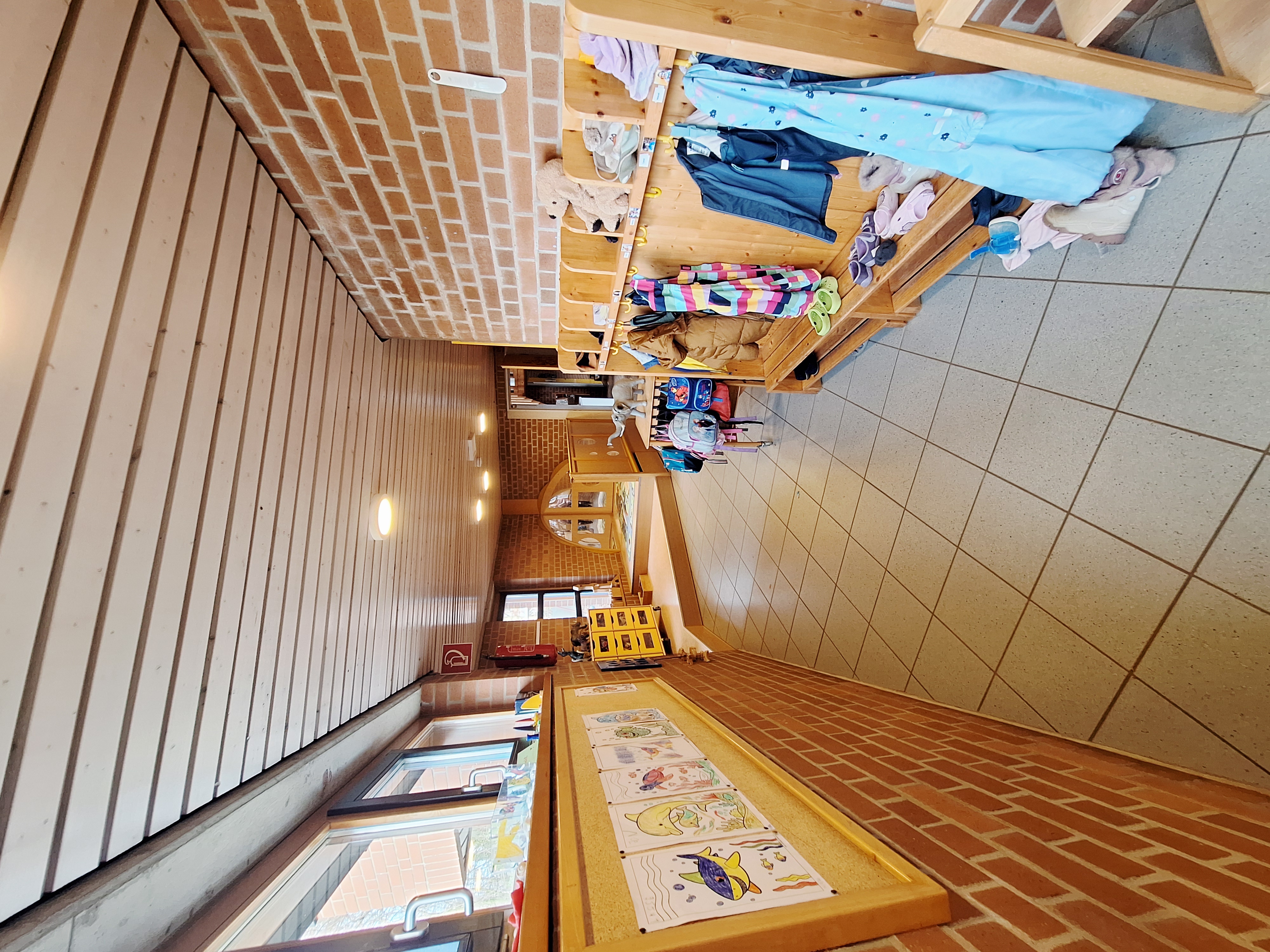 Hier sieht man den Flurbereich der Kita. Entlang der rechten Seite befindet sich Garderoben mit Hacken, an denen Jacken und Taschen der Kinder hängen. Der Flur führt weiter in die Gruppenräum