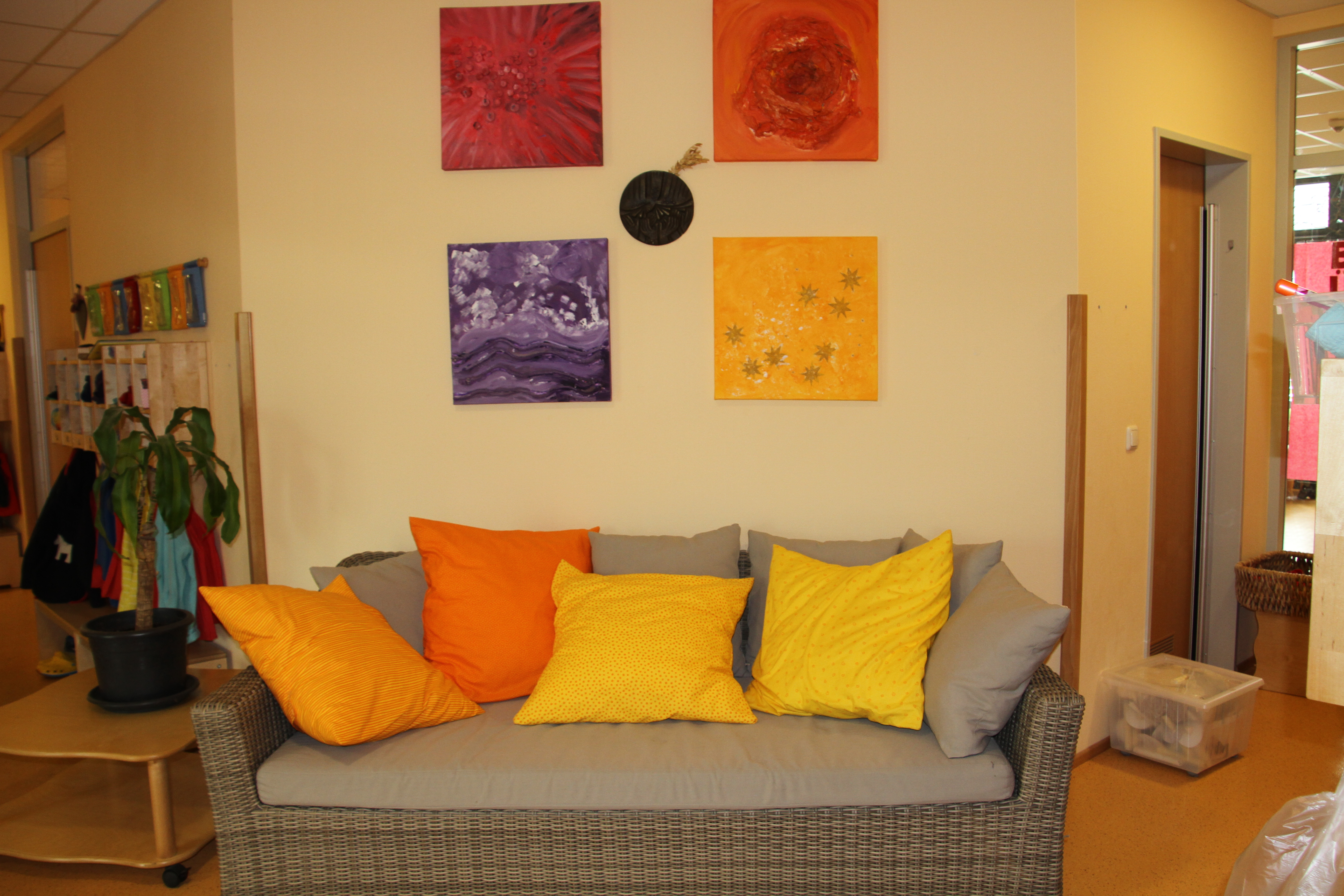 Das Foto zeigt den Eingangsbereich der Kita mit einem  großen grauen Korbmöbelsofa vor einer vanillefarbig gestrichenen Wand bestückt mit mehreren großen farbigen Kissen in orange- gelb Tönen