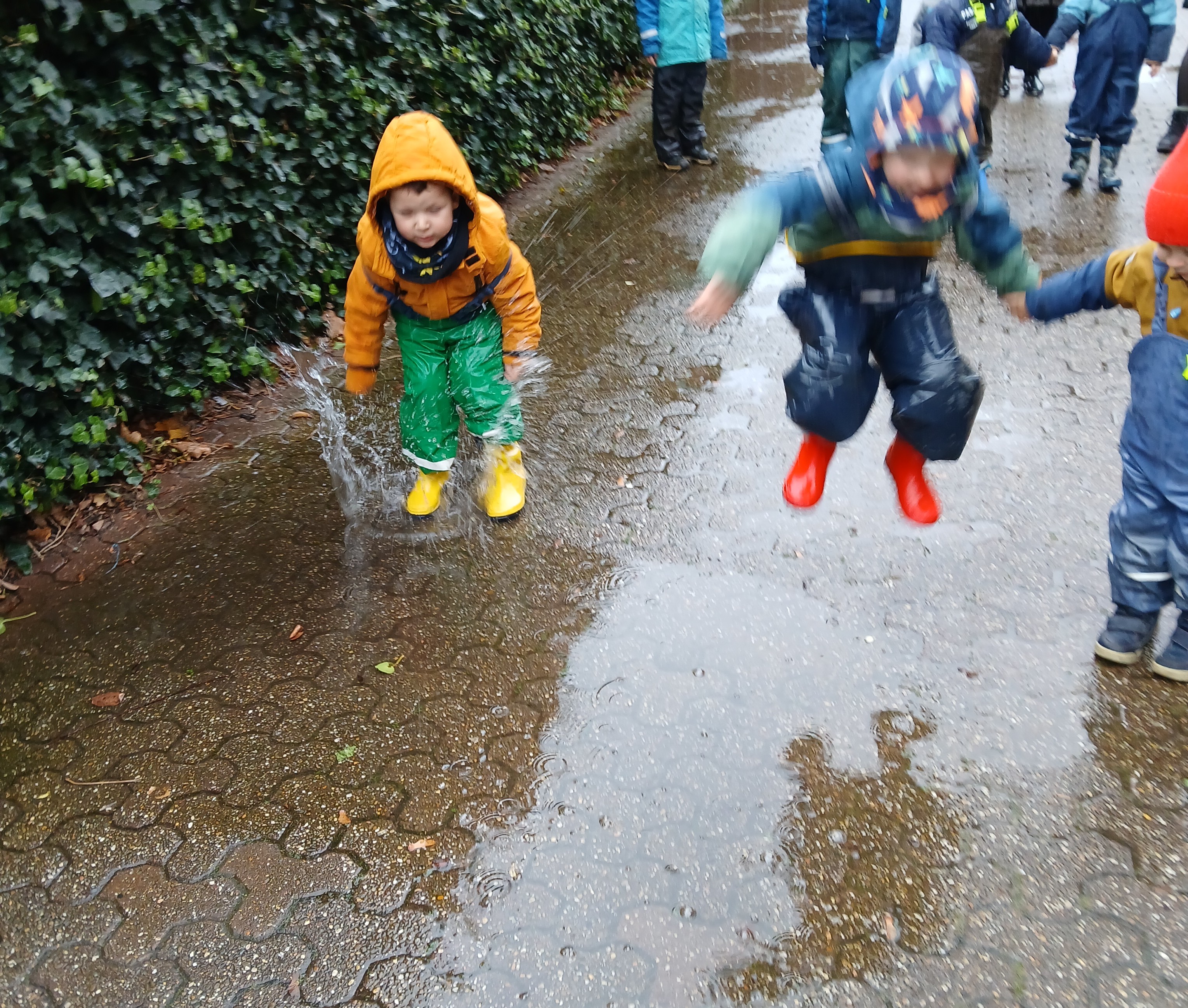 eine Gruppe von Kindern kommen von einem Ausflug zurück. Es ist Regenwetter und sie springen mit Begeisterung in Pfützen