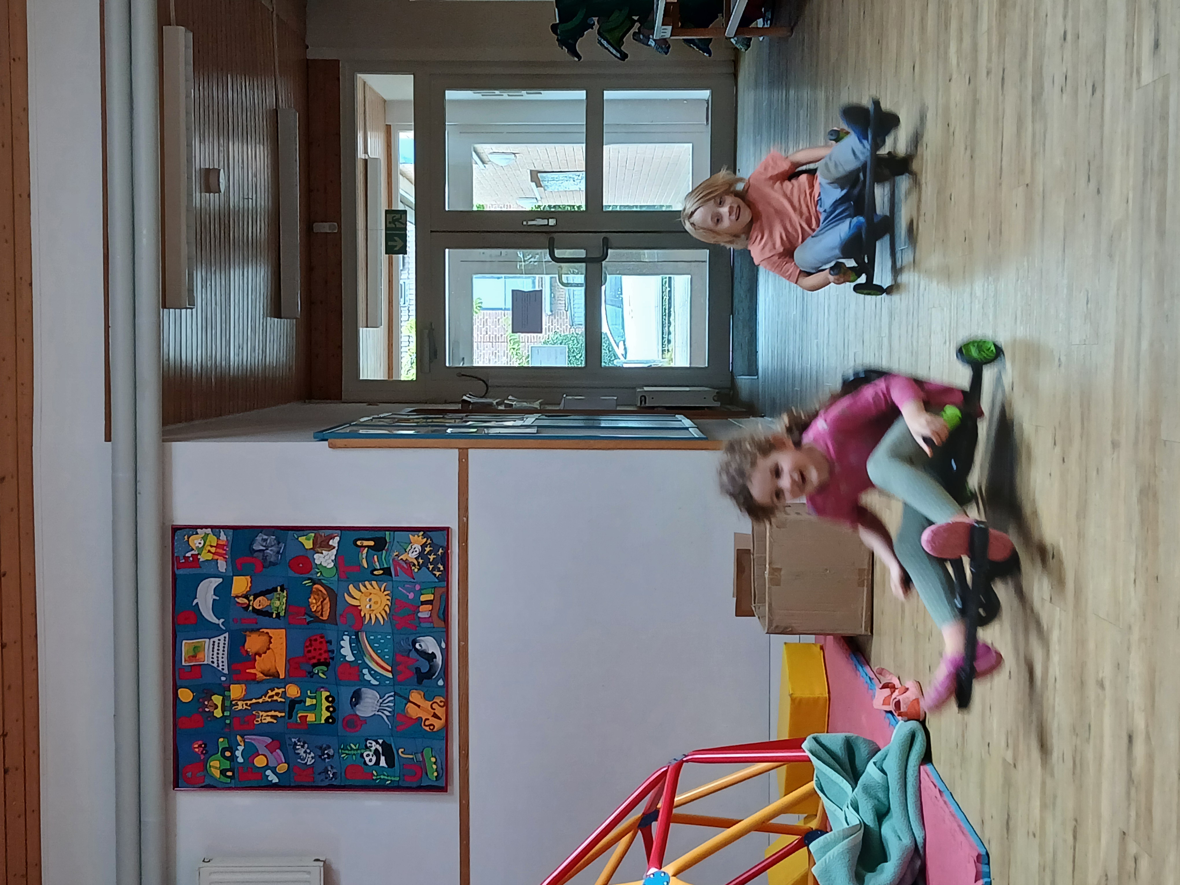 2 Kinder fahren mit Fahrzeugen in der Eingangshalle der Kita. Man erkennt, dass sie schnell unterwegs sind