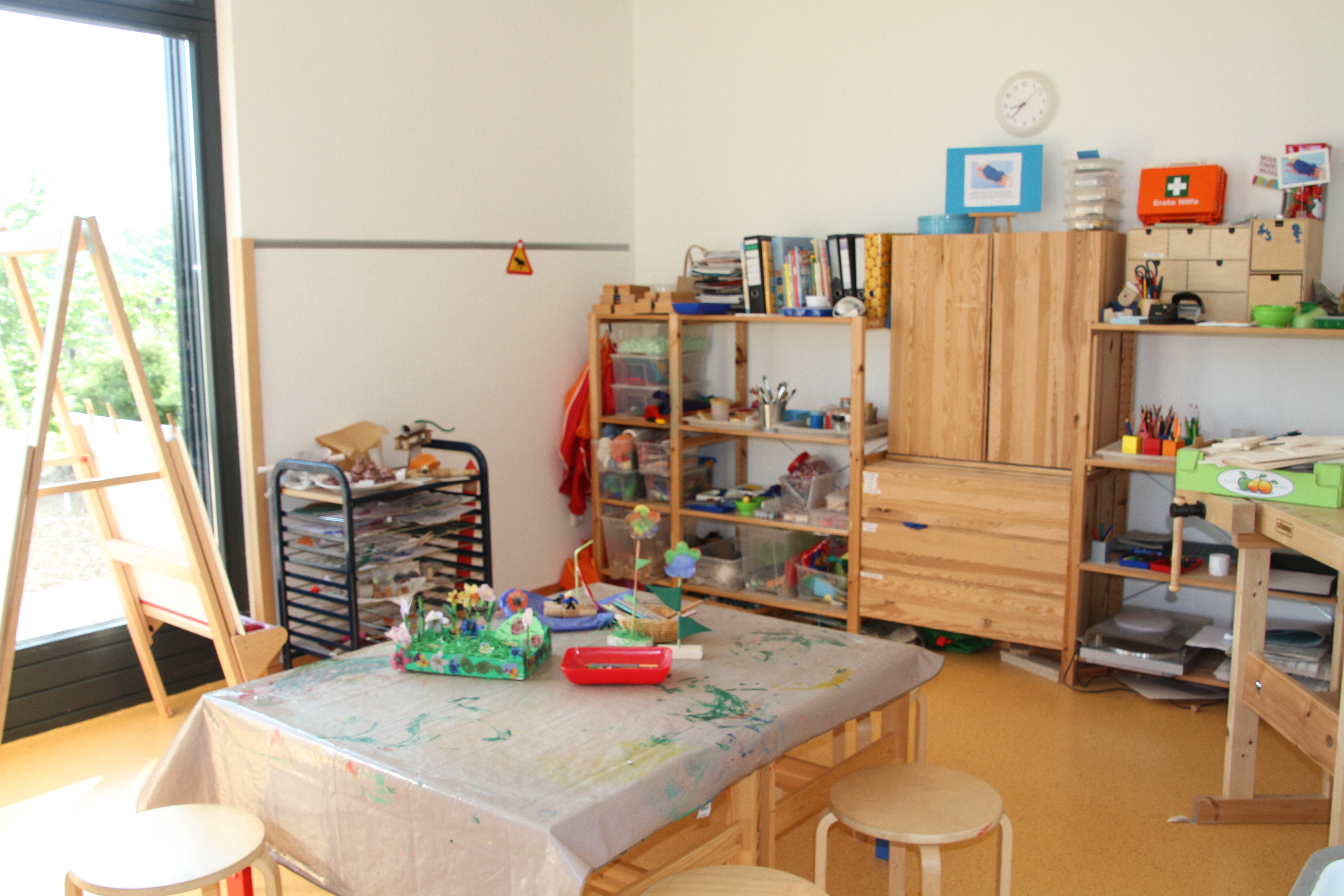 Das Bild zeigt einen weiß gestrichenen Raum mit bodentiefen Fensterelementen. In der mit ist eine Tischgruppe zu sehen, an der bis zu acht Kinder Platz nehmen können. Um die Tischgruppe stehe